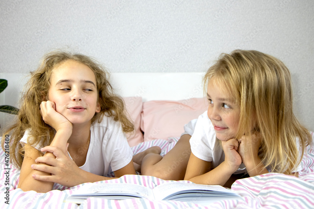 two cute sisters are lying on the bed and reading a funny book, smiling and laughing, family evening, one looking at the other, children's development through independent reading