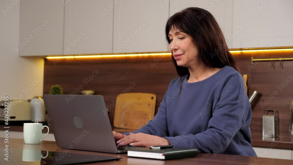 Smiling businesswoman works for laptop online chatting with company employees at home, distance meeting. Positive aged woman uses computer typing text on keyboard, sitting on chair at kitchen table Smiling businesswoman works for laptop online chatting with company employees at home, distance meeting. Positive aged woman uses computer typing text on keyboard, sitting on chair at kitchen table