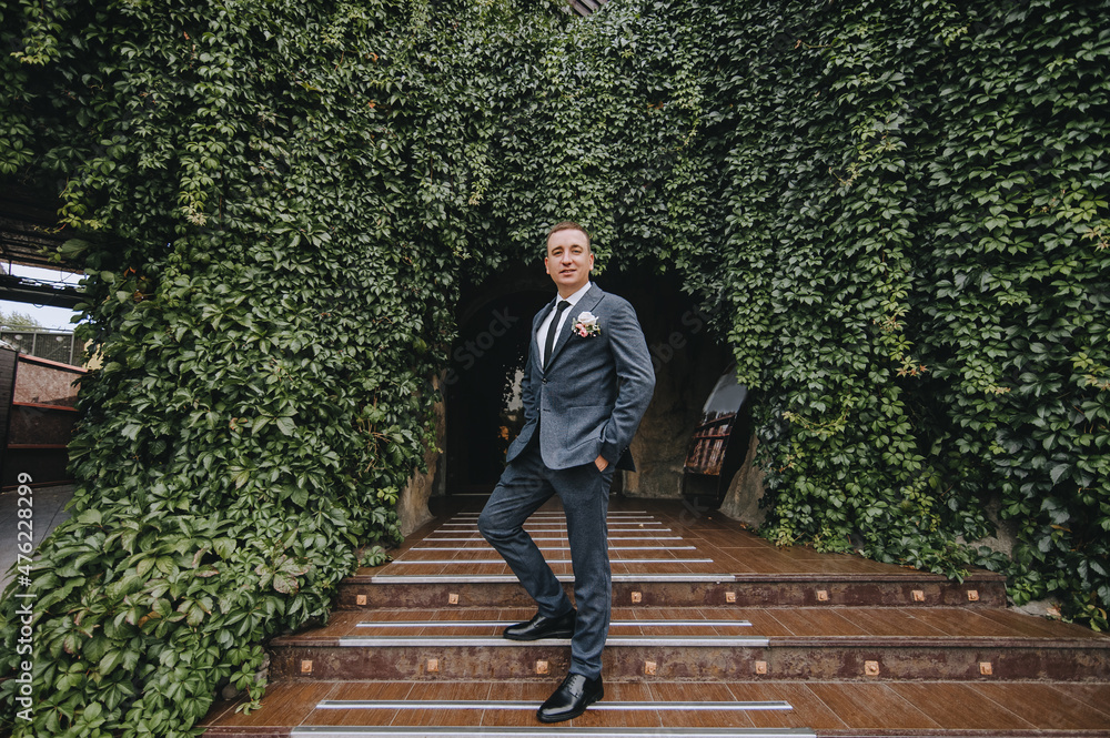 Fototapeta premium Stylish groom in a gray suit stands on the steps in nature, against the backdrop of green ivy foliage. Wedding portrait.