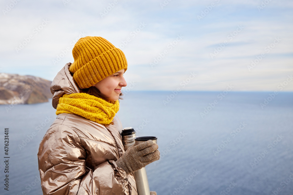 A young beautiful girl on a journey admires the beauty of nature. He holds a cup of hot tea from a thermos in his hands. Freedom, the concept of travel, active recreation.