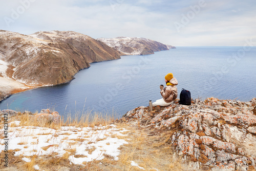 A young beautiful traveler girl is sitting on a rock overlooking Lake Baikal, holding a cup of hot tea from a thermos in her hand. Mountain winter landscape.