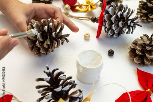 Handmade toy for the Christmas tree from pine cones. Children's creativity. Hands close-up paint the cone with white paint.
