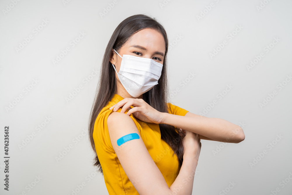 Portrait of health care asian young woman wearing face mask protect, getting vaccine of covid-19, influenza in yellow t-shirt, showing arm on blue bandage, isolated on nature background, copy space. Portrait of health care asian young woman wearing face mask protect, getting vaccine of covid-19, influenza in yellow t-shirt, showing arm on blue bandage, isolated on nature background, copy space.