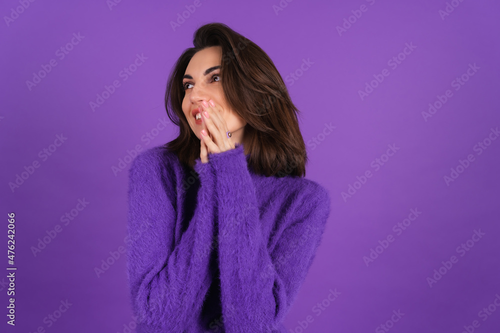 Fototapeta premium Young woman in a purple soft cozy sweater on the background is cute, in high spirits, laughs shyly, covering her mouth with her hands