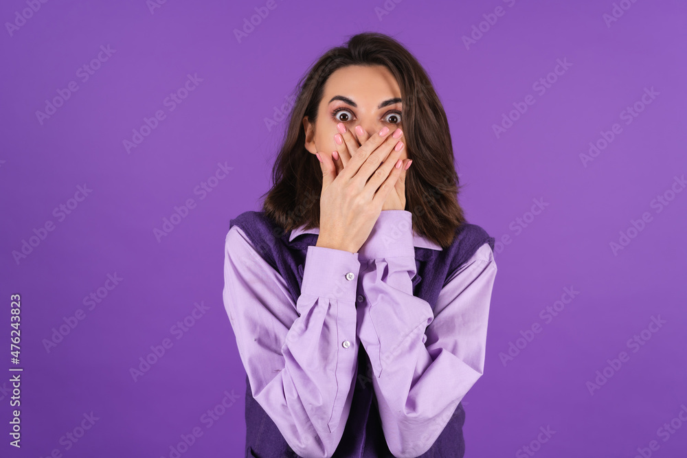 Young woman in a shirt and a vest on a purple background surprised shocked with an open mouth looks at the camera covers her mouth with her hands Young woman in a shirt and a vest on a purple background surprised shocked with an open mouth looks at the camera covers her mouth with her hands