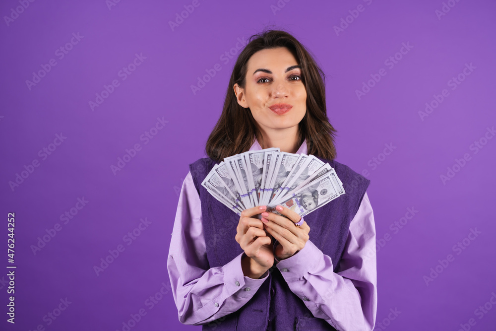 Young woman in a shirt and vest on a purple background with a bundle of money dollars in her hand smiles confidently Young woman in a shirt and vest on a purple background with a bundle of money dollars in her hand smiles confidently