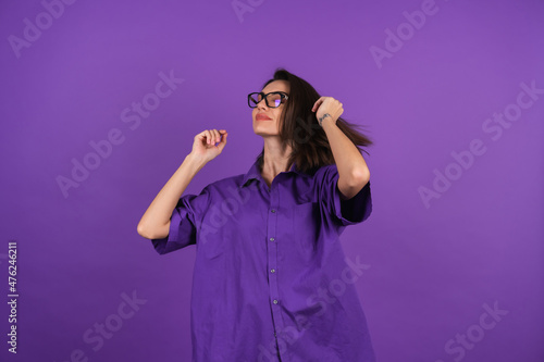 A young woman in a shirt with a short sleeve on a purple background, with makeup and stylish glasses, with closed eyes, enjoys, her hair is flying