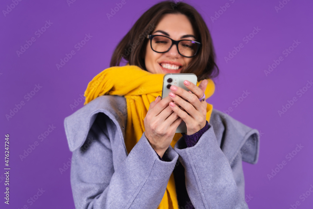Young woman in a gray coat, knitted dress and in a yellow scarf on a purple background, fashionable glasses, looks at the phone screen and laughs cheerfully Young woman in a gray coat, knitted dress and in a yellow scarf on a purple background, fashionable glasses, looks at the phone screen and laughs cheerfully