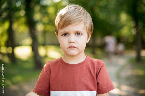 Little serious boy. It stands on a background of green park. The boy looks straight into the camera lens