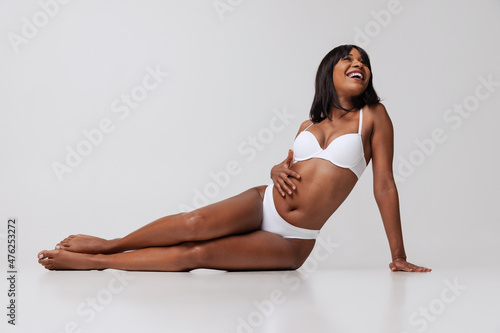 Young beautiful dark skinned girl in white underwear sitting on floor isolated over gray studio background. Natural beauty concept.