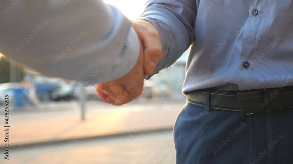 Dolly shot of two young colleagues meeting and shaking hands at city street on sunset. Successful businessmen greeting each other against the background of cars parking. Business handshake outdoor Dolly shot of two young colleagues meeting and shaking hands at city street on sunset. Successful businessmen greeting each other against the background of cars parking. Business handshake outdoor