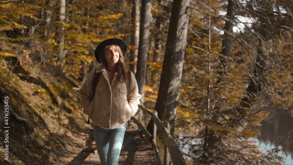 Woman tourist walks on trail in fall forest on sunny autumn day. female hiker in hat with backpack hiking in forest around mountain lake at cold sunny morning, adventure outdoor lifestyle. Woman tourist walks on trail in fall forest on sunny autumn day. female hiker in hat with backpack hiking in forest around mountain lake at cold sunny morning, adventure outdoor lifestyle.