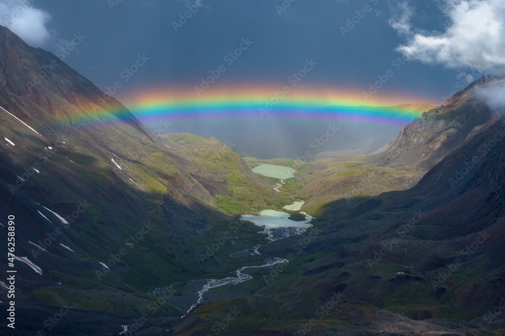 Rainbow over a mountain valley. Atmospheric alpine landscape with snowy mountains with rainbow in rainy and sunny weather.