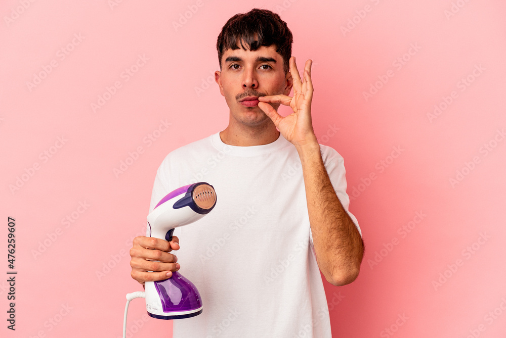 Young mixed race man holding an iron isolated on pink background with fingers on lips keeping a secret.