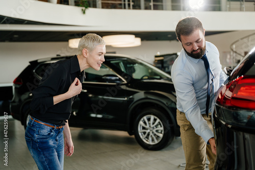 Positive, friendly car seller showing to attentive female customer in black shirt how to open the trunk from the outside. People in a auto salon or at showroom. Concept of people and transport. © Iryna