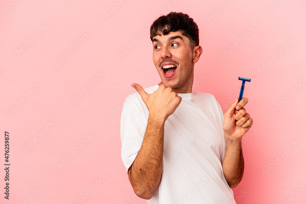 Young mixed race man holding razor blade isolated on pink background points with thumb finger away, laughing and carefree. Young mixed race man holding razor blade isolated on pink background points with thumb finger away, laughing and carefree.