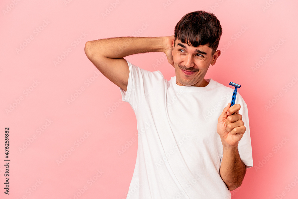 Young mixed race man holding razor blade isolated on pink background touching back of head, thinking and making a choice. Young mixed race man holding razor blade isolated on pink background touching back of head, thinking and making a choice.