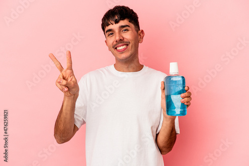 Young mixed race man holding mouthwash isolated on pink background joyful and carefree showing a peace symbol with fingers.