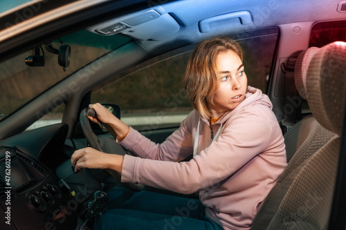Portrait of a frightened young Caucasian woman driving a right-handed car, looking back at the police siren light. Concepts of road accidents and chase photo