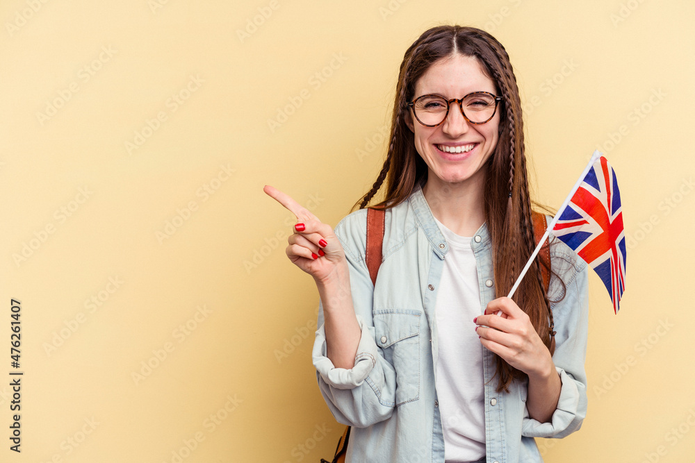 Young caucasian woman studying English isolated on yellow background smiling and pointing aside, showing something at blank space. Young caucasian woman studying English isolated on yellow background smiling and pointing aside, showing something at blank space.