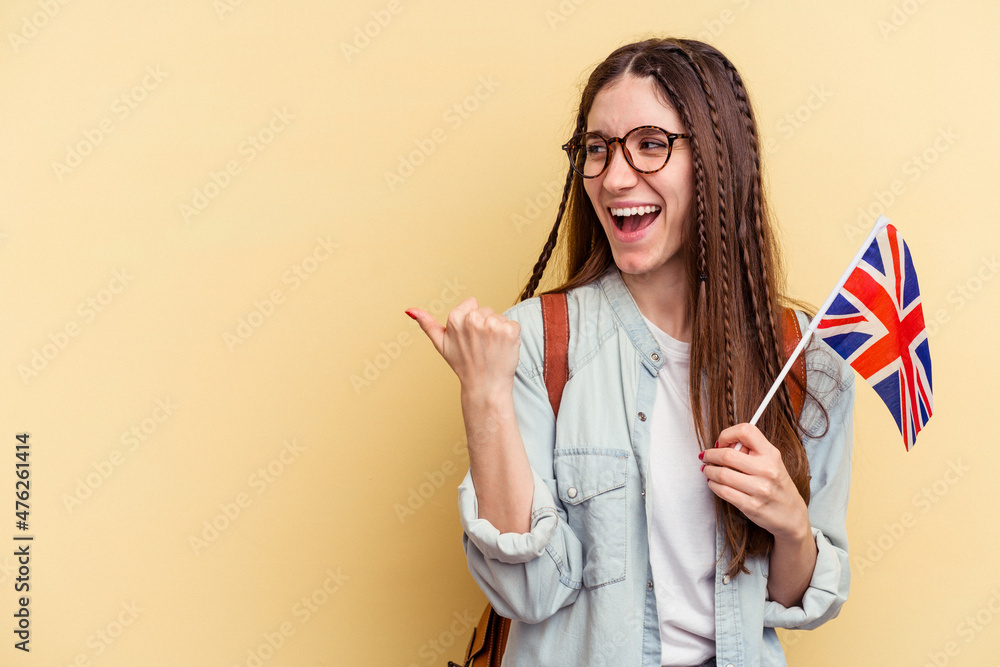 Young caucasian woman studying English isolated on yellow background points with thumb finger away, laughing and carefree.