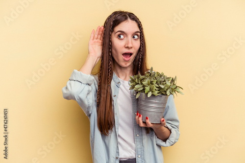 Young caucasian woman holding a plant isolated on yellow background trying to listening a gossip.