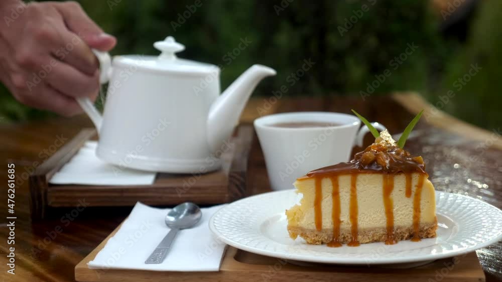 Man hand pouring hot tea in tea cup. Piece of delicious fresh cake on wooden table with white teapot in outdoor cafe. Drinking tea with cheesecake with caramel and macadamia nuts. Sweet food Man hand pouring hot tea in tea cup. Piece of delicious fresh cake on wooden table with white teapot in outdoor cafe. Drinking tea with cheesecake with caramel and macadamia nuts. Sweet food