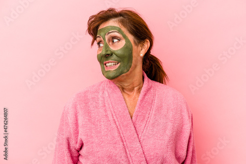 Middle age caucasian woman wearing a facial mask isolated on pink background looks aside smiling, cheerful and pleasant.