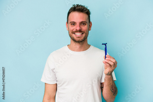 Young caucasian man shaving his beard isolated on blue background happy, smiling and cheerful.