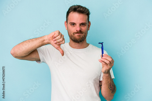 Young caucasian man shaving his beard isolated on blue background showing a dislike gesture, thumbs down. Disagreement concept.