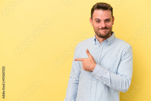 Young caucasian man isolated on yellow background smiling and pointing aside, showing something at blank space.