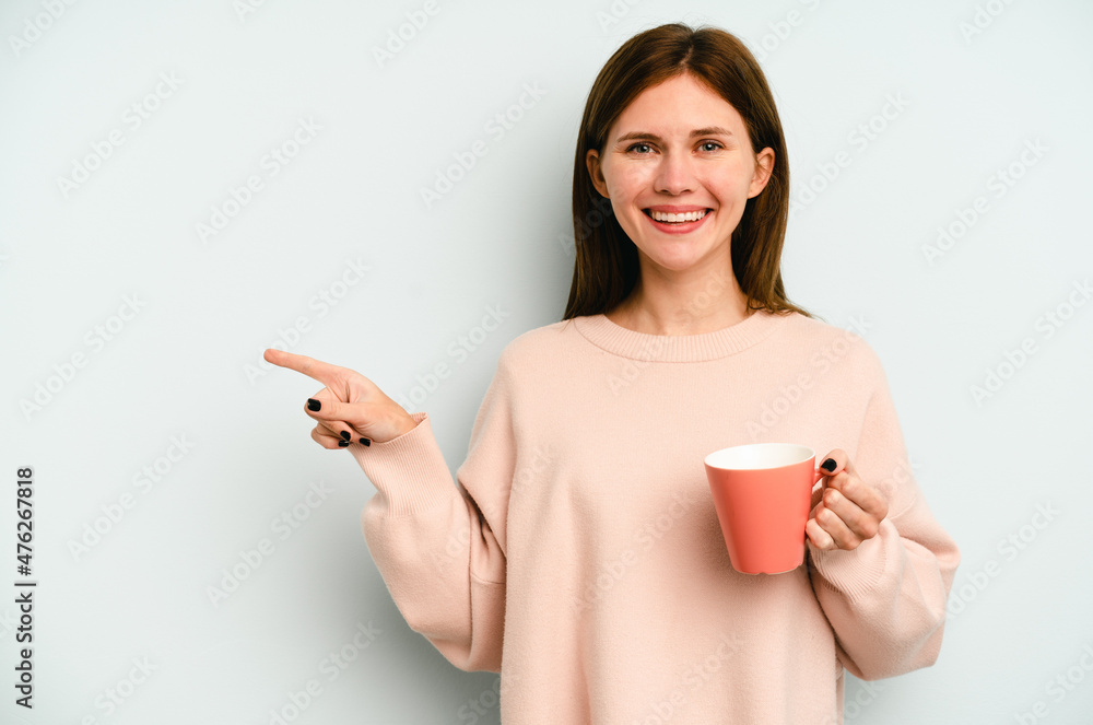 Young English woman holding a mug isolated on blue background smiling and pointing aside, showing something at blank space. Young English woman holding a mug isolated on blue background smiling and pointing aside, showing something at blank space.