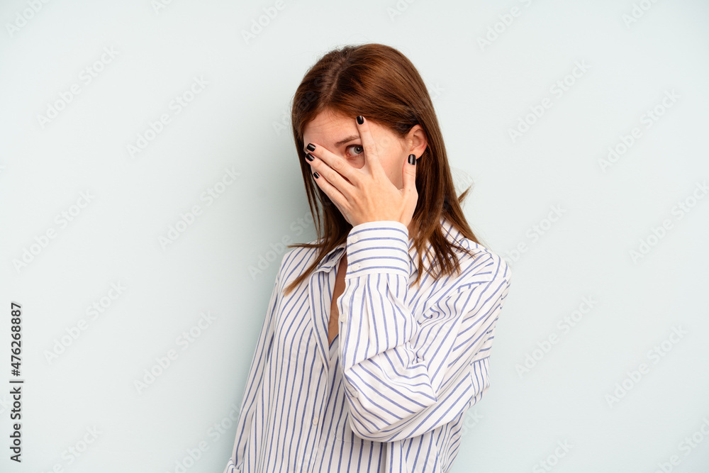 Young English woman isolated on blue background blink at the camera through fingers, embarrassed covering face. Young English woman isolated on blue background blink at the camera through fingers, embarrassed covering face.