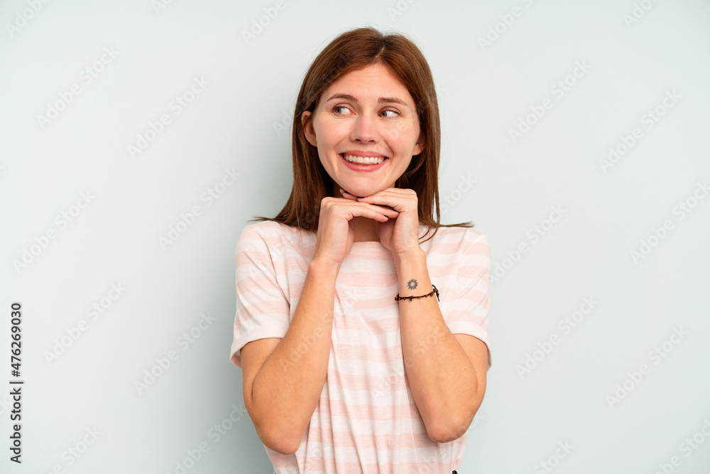 Young English woman isolated on blue background keeps hands under chin, is looking happily aside. Young English woman isolated on blue background keeps hands under chin, is looking happily aside.
