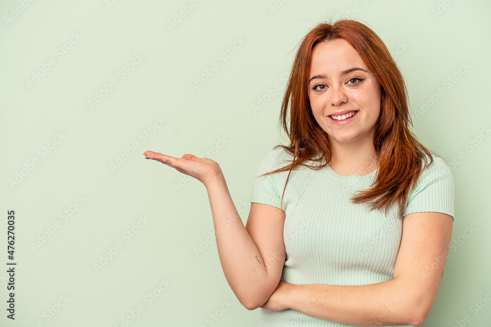 Young caucasian woman isolated on green background showing a copy space on a palm and holding another hand on waist. Young caucasian woman isolated on green background showing a copy space on a palm and holding another hand on waist.
