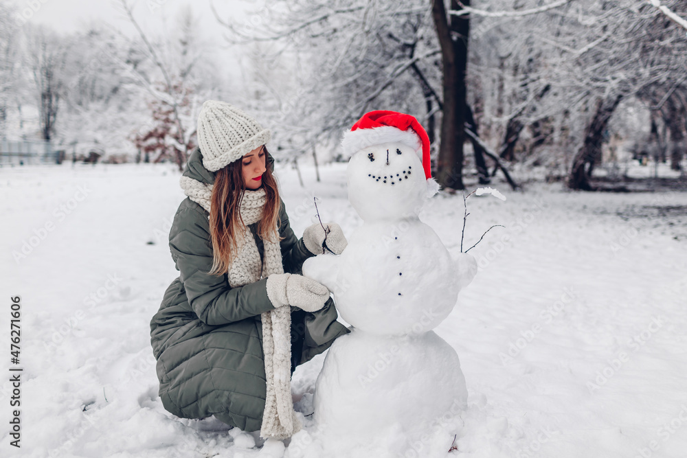 Girl makes snowman outdoors in snowy winter park dressing it in Santa hat, scarf and attaching branches in hands. Girl makes snowman outdoors in snowy winter park dressing it in Santa hat, scarf and attaching branches in hands.