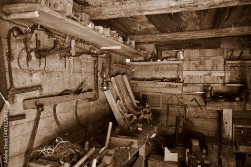 A sepia photo of a medieval workshop including a set of tools for processing wood, metal, farming tools, an oil lamp and other useful equipment seen on a Polish countryside during summer