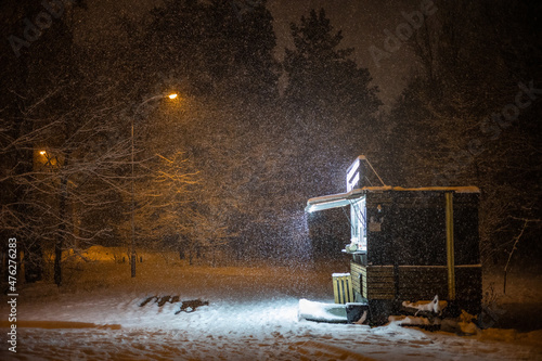 A lonely coffee stall that is brightly lit in a dark remote corner of a large city park during an evening snowfall on Christmas Eve.