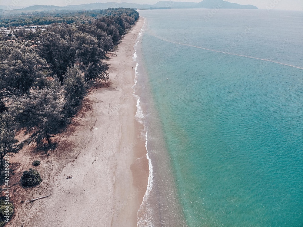Aerial photography made by drone of a tropical and exotic seascape. The sandy kata beach is washed with white waves. Light warm sand with standing green trees, developing in the wind.
