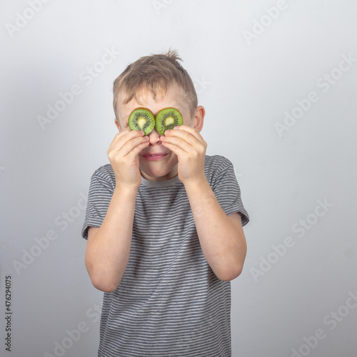 A cheerful Caucasian boy closed his eyes with slices of green kiwi fruit on a gray background in the studio