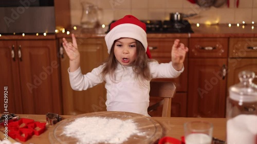 Adorable little girl in red santa hat dancind by the table. Preparing for Christmas at home. Baking gingerbread photo