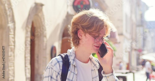 Cheerful Caucasian male teenager talking to family on cellphone in city. Young man guy wearing casual clothes in glasses, using smartphone outdoors. Technology, youth, education concept. photo