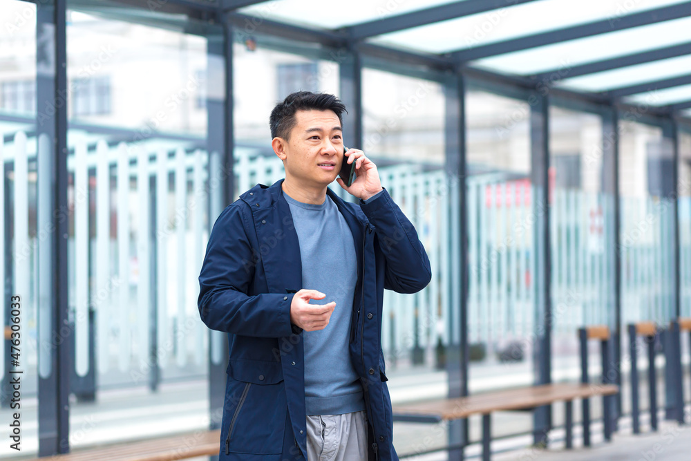 A man talking on the phone, focused and serious Asian at a public transport stop, a businessman near the office center A man talking on the phone, focused and serious Asian at a public transport stop, a businessman near the office center