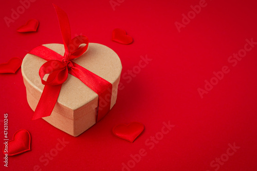 Beautiful minimal wrapped gift box heart shaped with red bow ribbon on red background and small padded hearts top view flat lay, present for Saint Valentine day, love and relationship, space for text