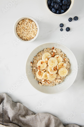 Bowl of porridge topped with banana and almonds,  a bowl of oats and a  bowl of blueberries on white marble countertop