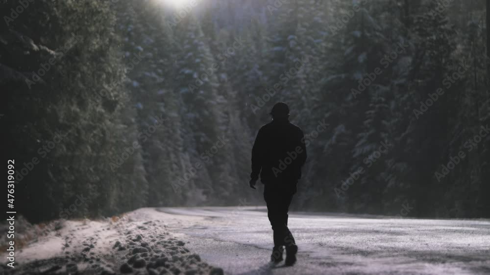A silhouetted man walks down a snowy road leading into tall evergreens as the sun shines down from above. A silhouetted man walks down a snowy road leading into tall evergreens as the sun shines down from above.