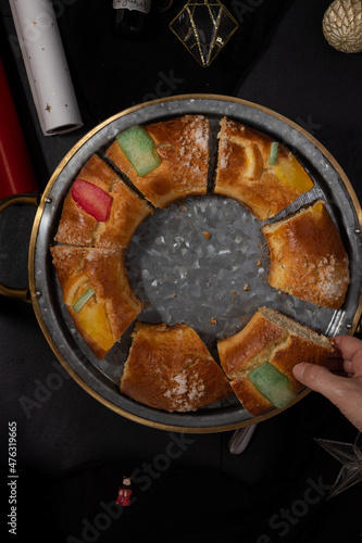 Woman right hand taking a just cut sweet piece of King cake or spanish Roscón de Reyes from a metallic tray over a dark and elegant background.