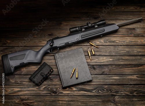 A modern bolt-action scout rifle with a telescopic sight. Weapon on a wooden background. Cartridges and a black book