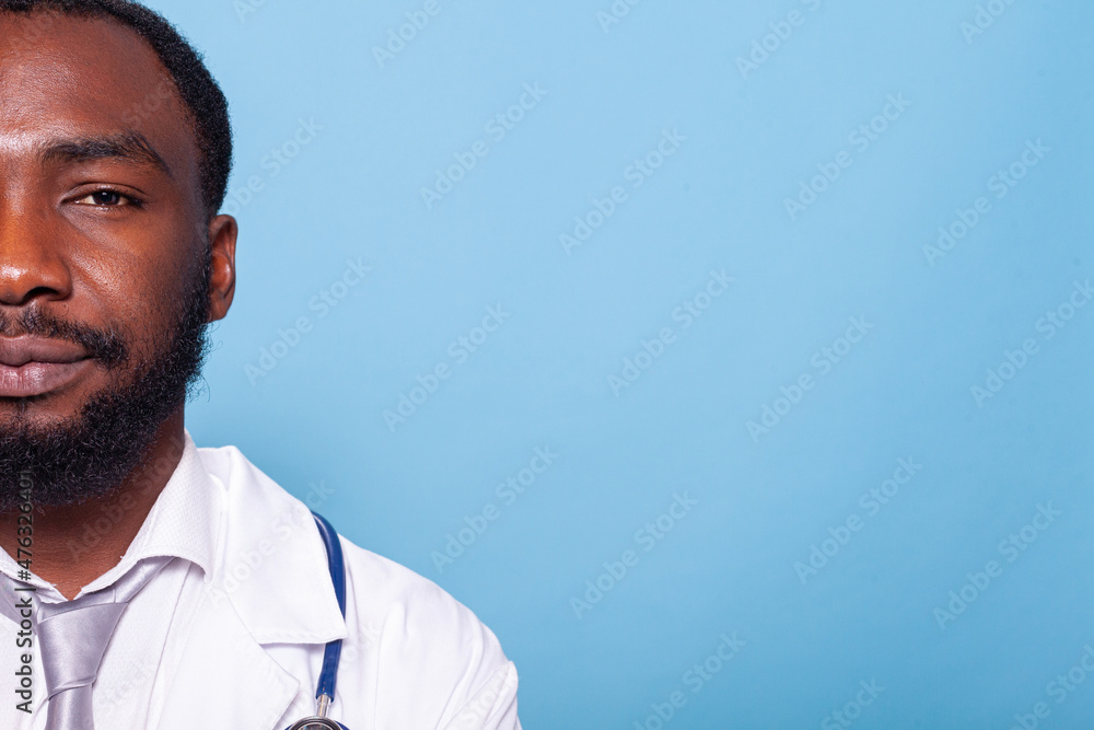 Half face closeup portrait of smiling medical doctor in white lab coat with stethoscope on blue background. Cropped image of african american medic looking confident and relaxed. Half face closeup portrait of smiling medical doctor in white lab coat with stethoscope on blue background. Cropped image of african american medic looking confident and relaxed.