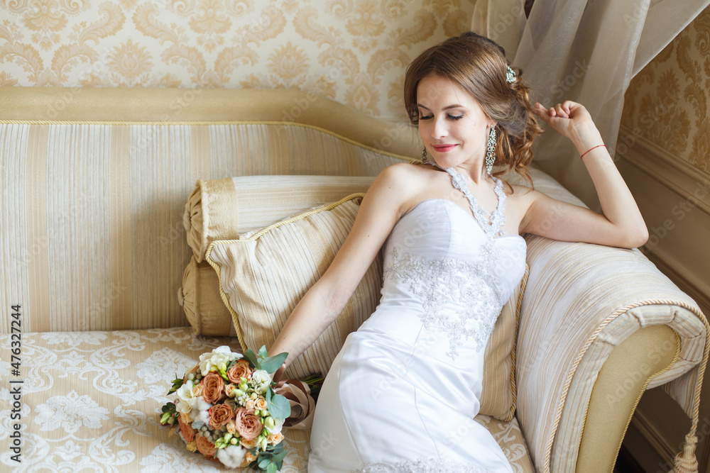 Beautiful young Bride with classic hairstyle sits on a sofa in a bedroom. Classic fish-silhouette white wedding dress. Wedding fashion